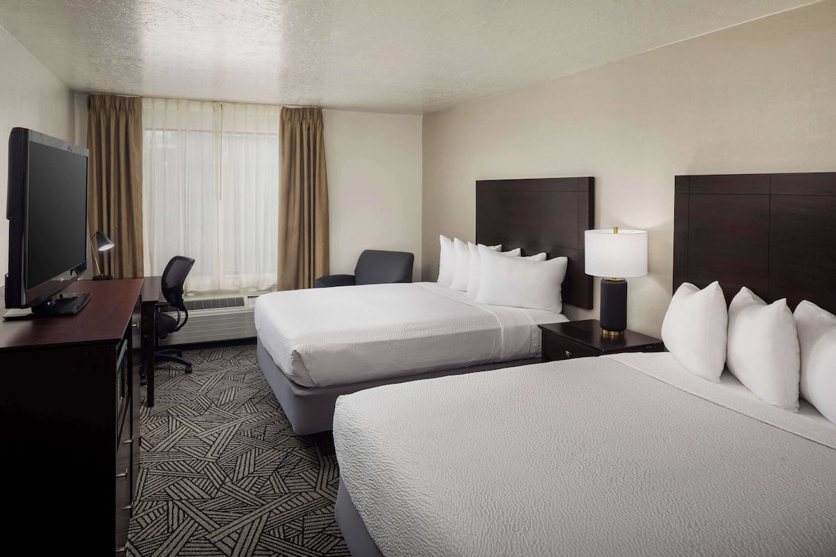 A hotel room with two beds, a desk with a TV, a chair, a lamp, and a large window with curtains.