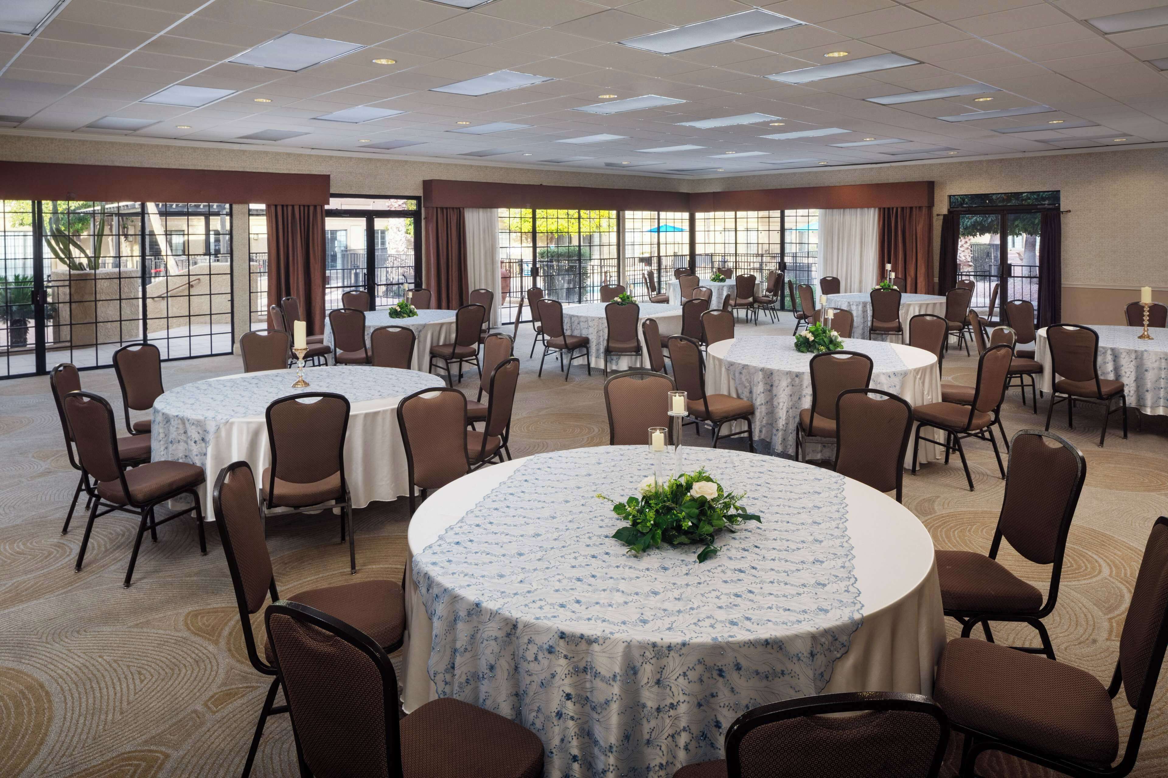 A large conference room with multiple round tables, each surrounded by brown chairs. The room has large windows with curtains, allowing natural light to enter.