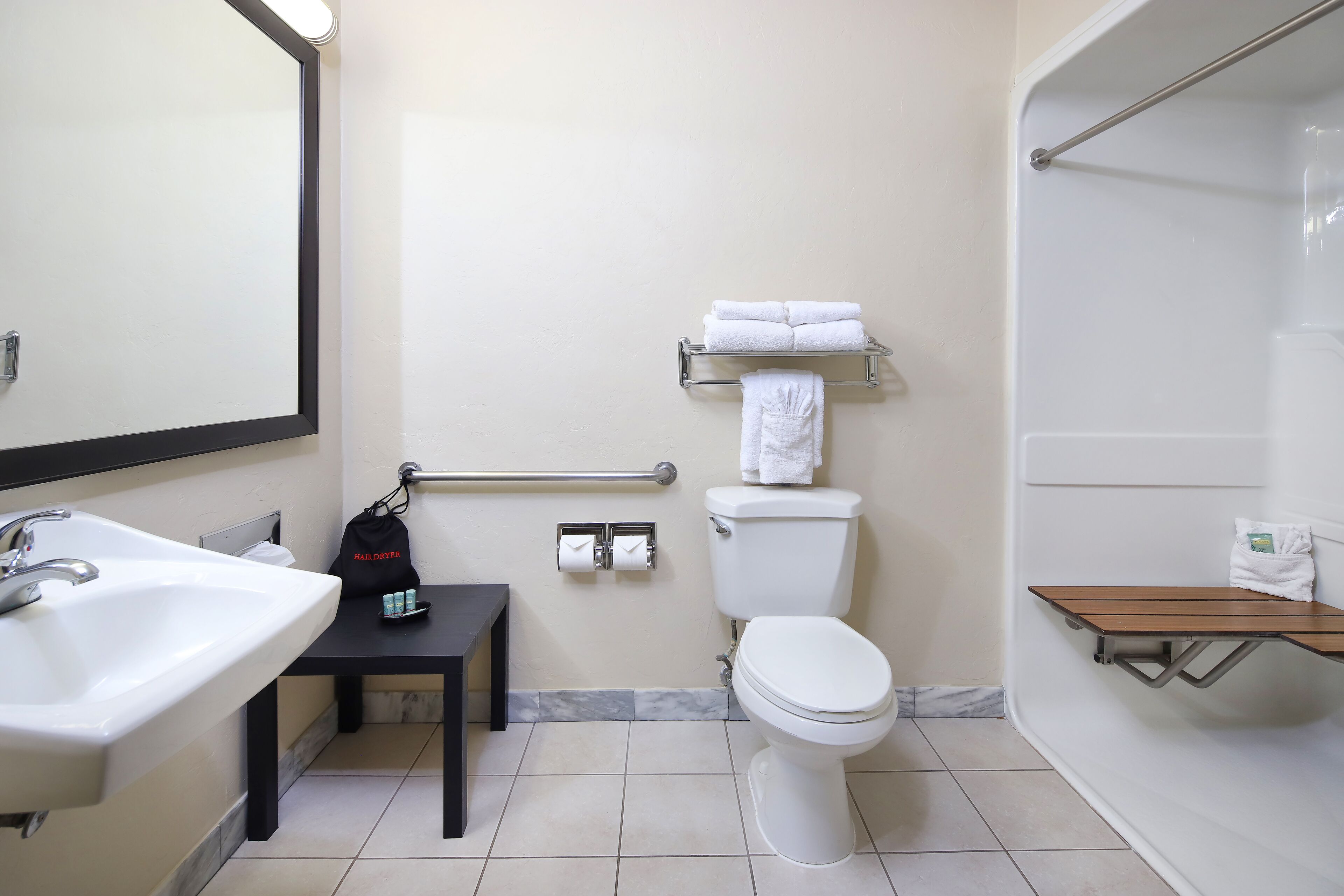 A bathroom with a sink, toilet, and shower. There is a towel rack with towels, a small table with a bag, and a shelf with toiletries.