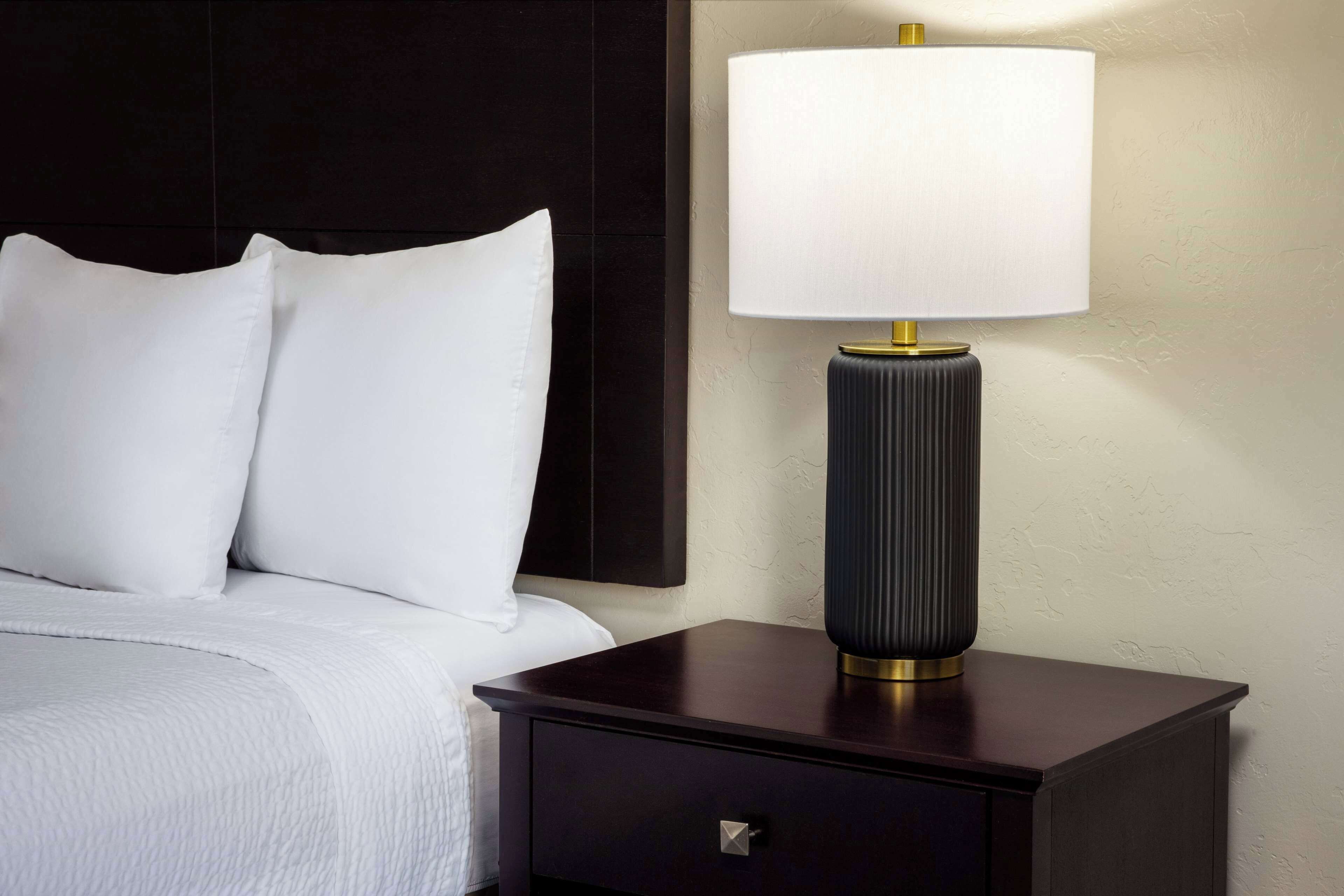 A neatly made bed with white linens and pillows, a dark wooden nightstand with a lamp, and a wall-mounted panel.