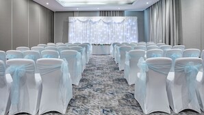 Une salle de conférence avec des rangées de chaises blanches ornées de rubans bleu clair, un sol recouvert de moquette et un plafond doté d’un éclairage encastré.