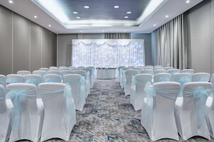 Un salón de conferencias con filas de sillas blancas con cintas azul claro, suelo alfombrado e iluminación empotrada en el techo.