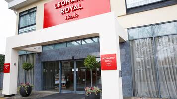 Exterior del Hotel Leonardo Royal con un llamativo letrero rojo, amplios ventanales de vidrio y dos plantas en macetas en la entrada.