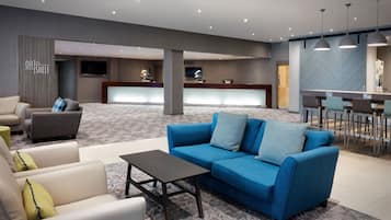 Un moderno vestíbulo de hotel con una recepción, una sala de estar con sofás y sillones, y un bar con taburetes.