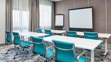 A conference room with a white table, teal chairs, a projector screen, and a large window with curtains.