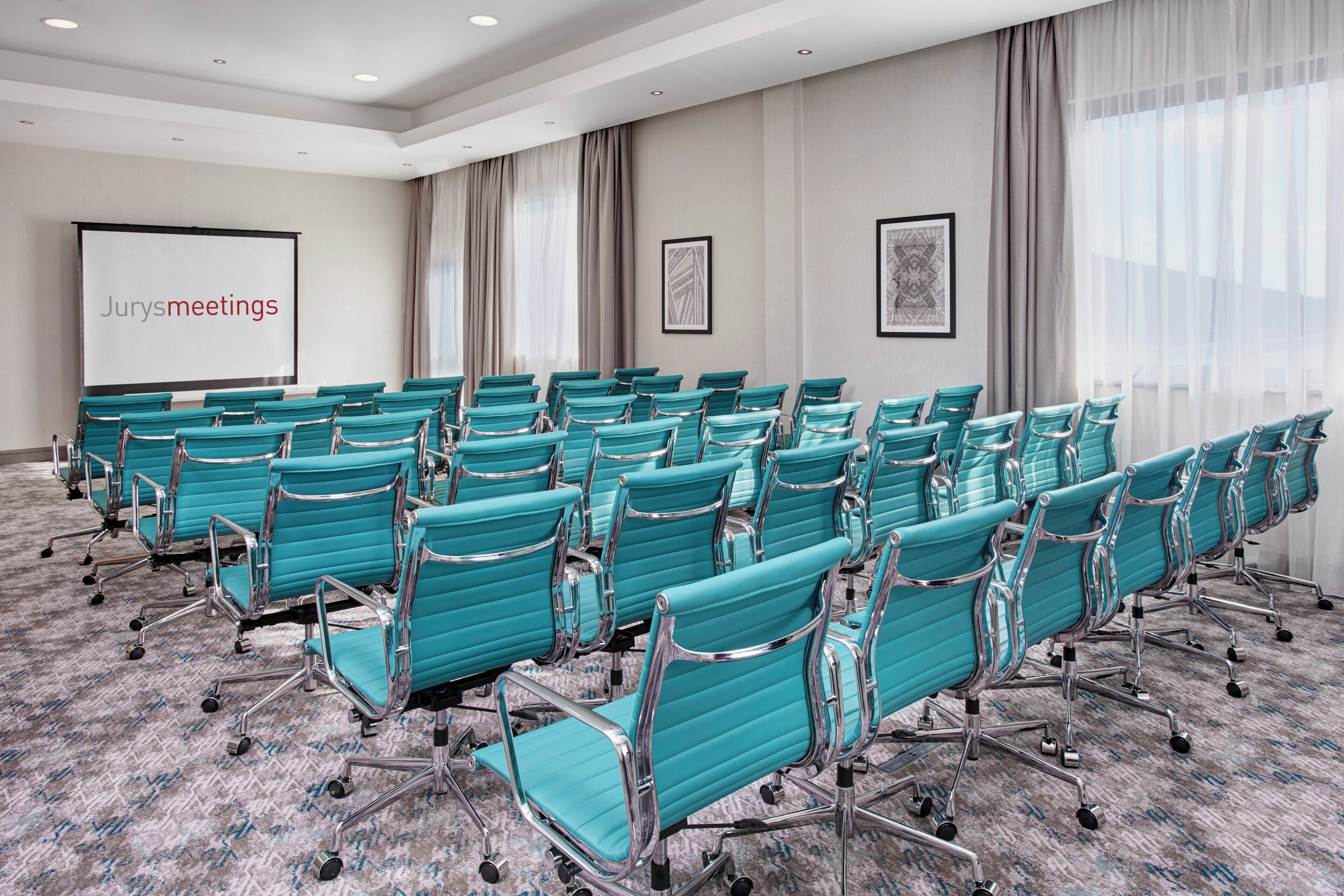Een vergaderruimte met rijen smaragdgroene stoelen, een scherm waarop 'Jurysmeetings' staat, en twee ingelijste kunstwerken aan de muren.