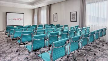 A conference room with rows of teal chairs, a projector screen with 'Jurysmeetings' text, and two framed artworks on the walls.