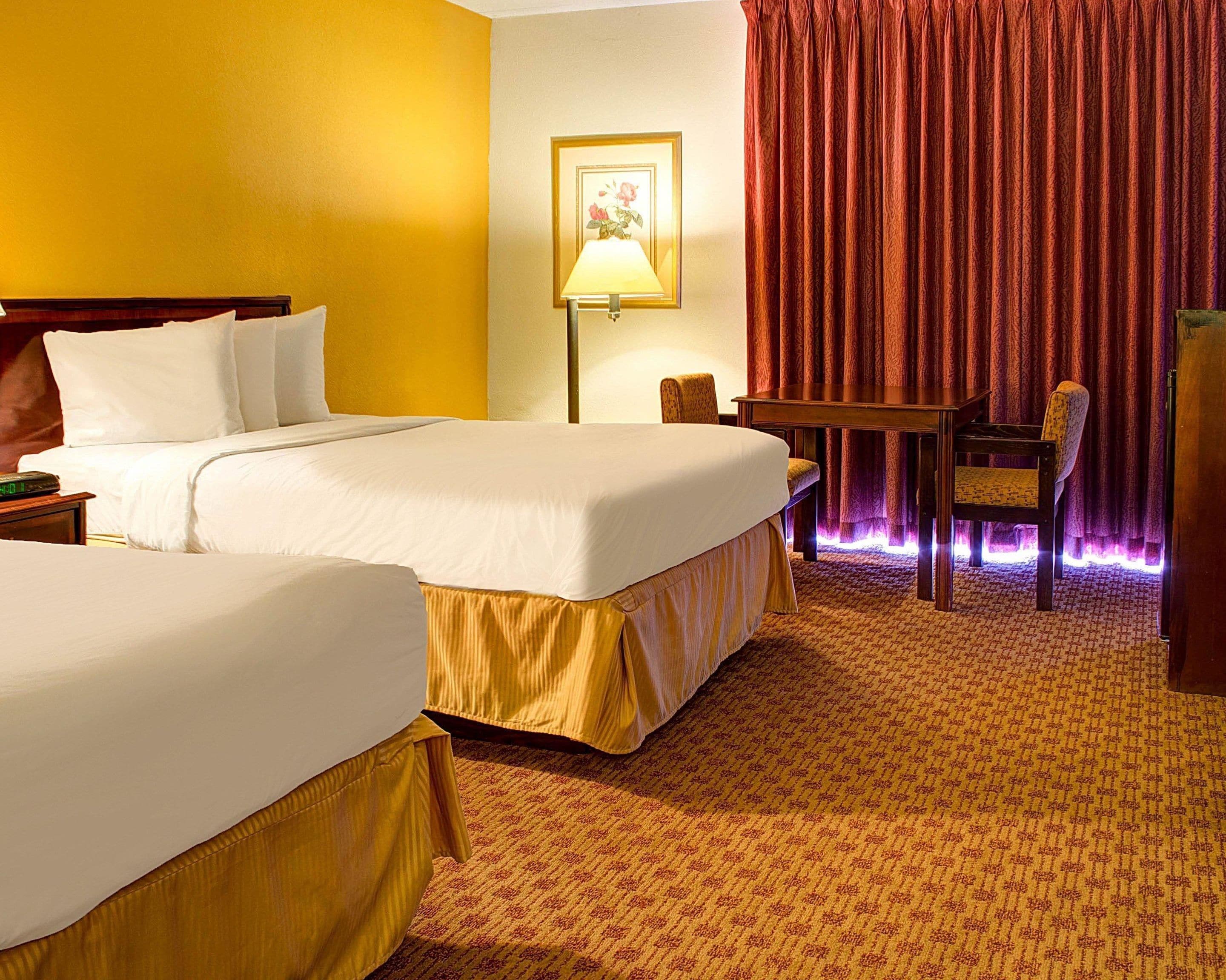 Ein Hotelzimmer mit zwei Betten, einem Schreibtisch, einem Stuhl, einer Lampe und einem gerahmten Bild an der Wand.