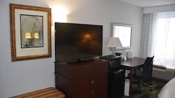 Ein Hotelzimmer mit einem großen Flachbildfernseher, einem Schreibtisch mit Lampe, einem Sessel, einer Bank und einem gerahmten Bild an der Wand.