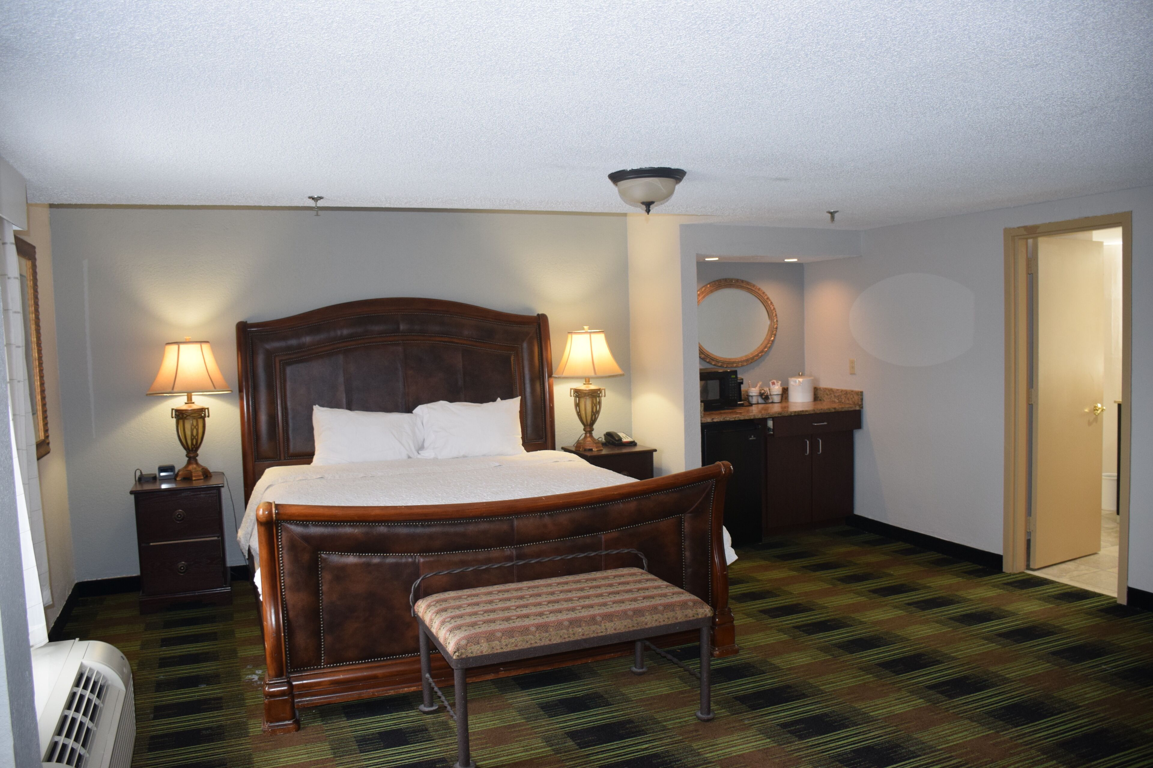 Habitación de hotel con una cama grande, lámparas de noche, un banco, un espejo, un escritorio y una puerta.