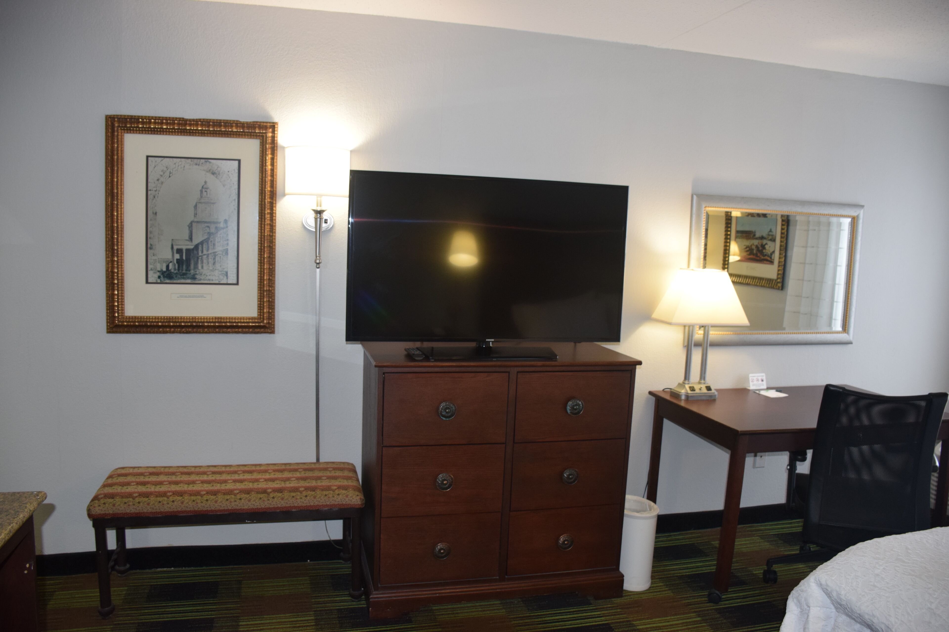 Une chambre d’hôtel avec une télévision posée sur une commode en bois, un banc, un bureau avec une chaise et un tableau encadré accroché au mur.