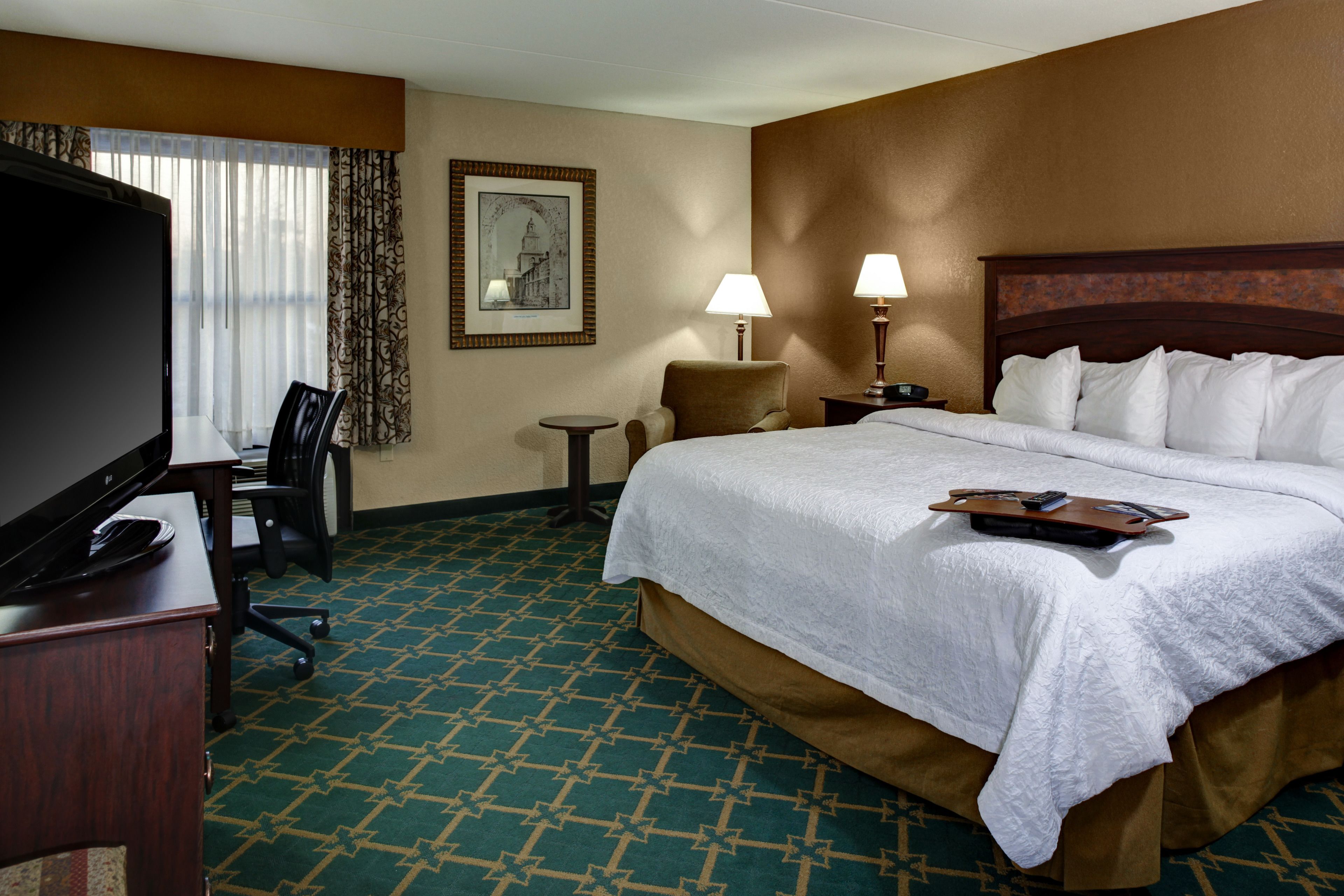 A hotel room with a large bed, a desk with a television, a chair, a small table, and a lamp.