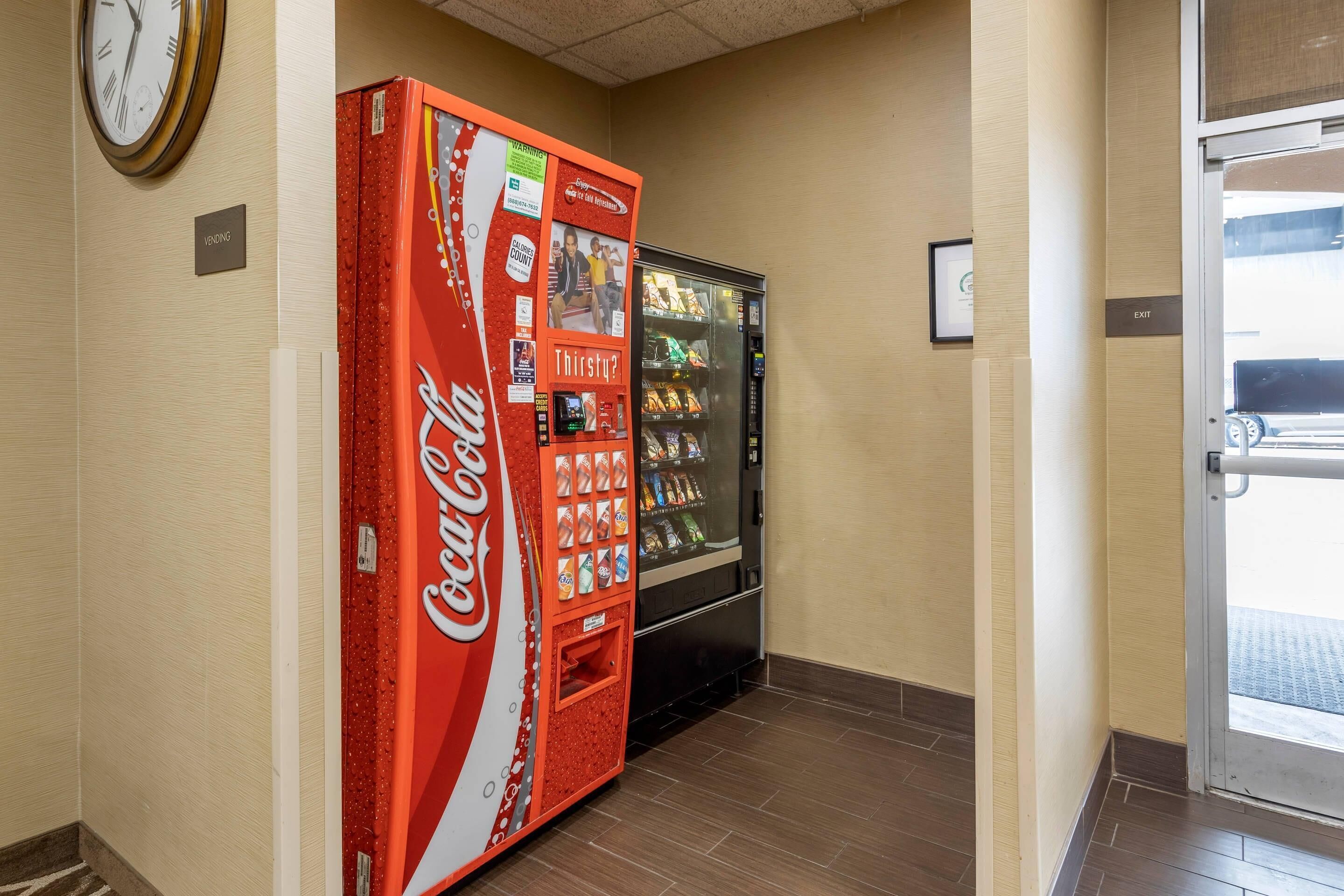 A vending machine and a Coca-Cola branded machine in a hallway with a clock on the wall and an exit sign.