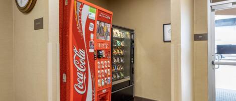 A vending machine and a Coca-Cola branded machine in a hallway with a clock on the wall and an exit sign.