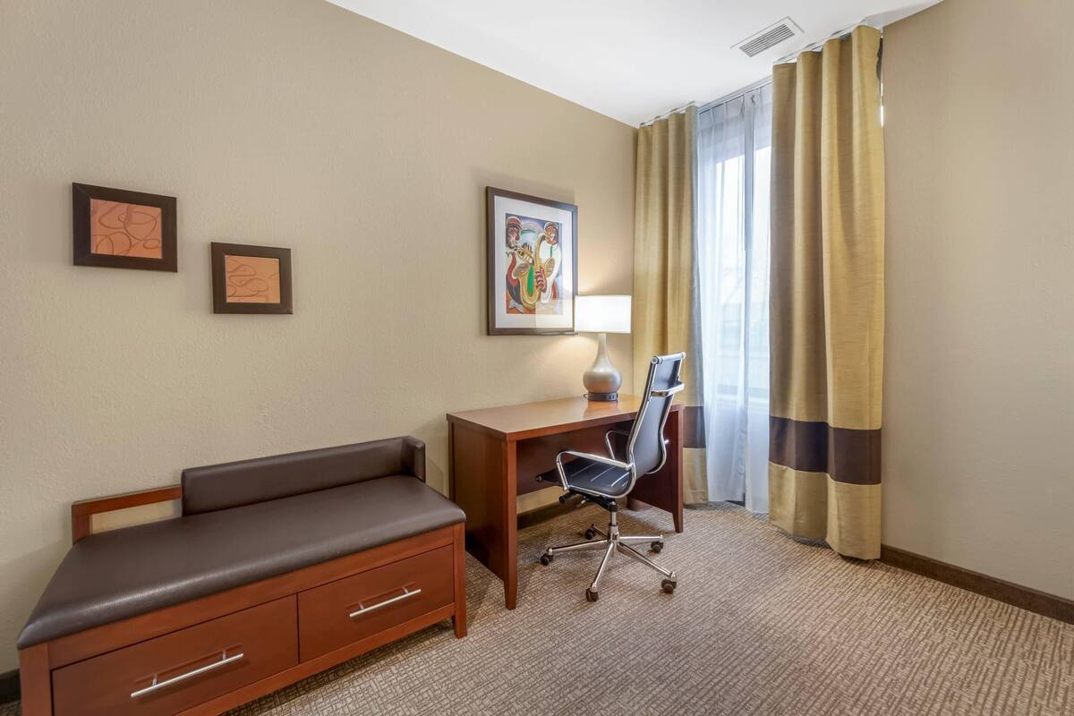 A hotel room with a wooden desk, a black office chair, a brown sofa, and two framed pictures on the wall.