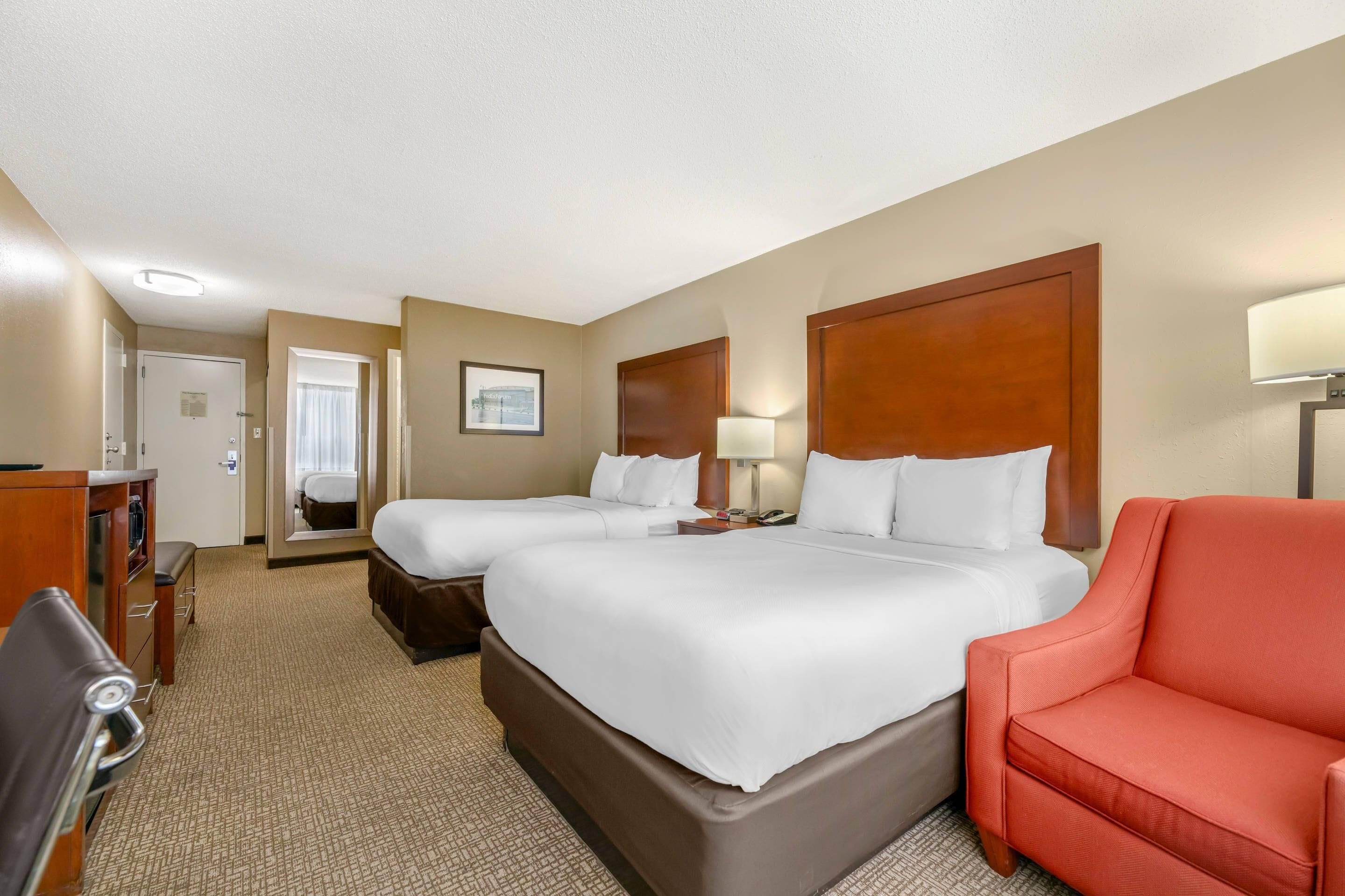 A hotel room with two beds, a desk, a chair, and a lamp.