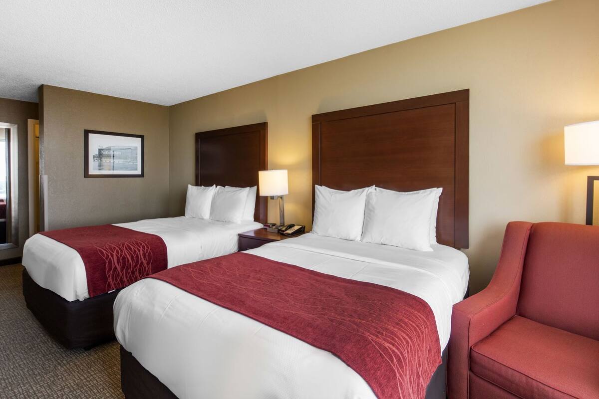 A hotel room with two beds, a red chair, a nightstand with a lamp, and a framed picture on the wall.