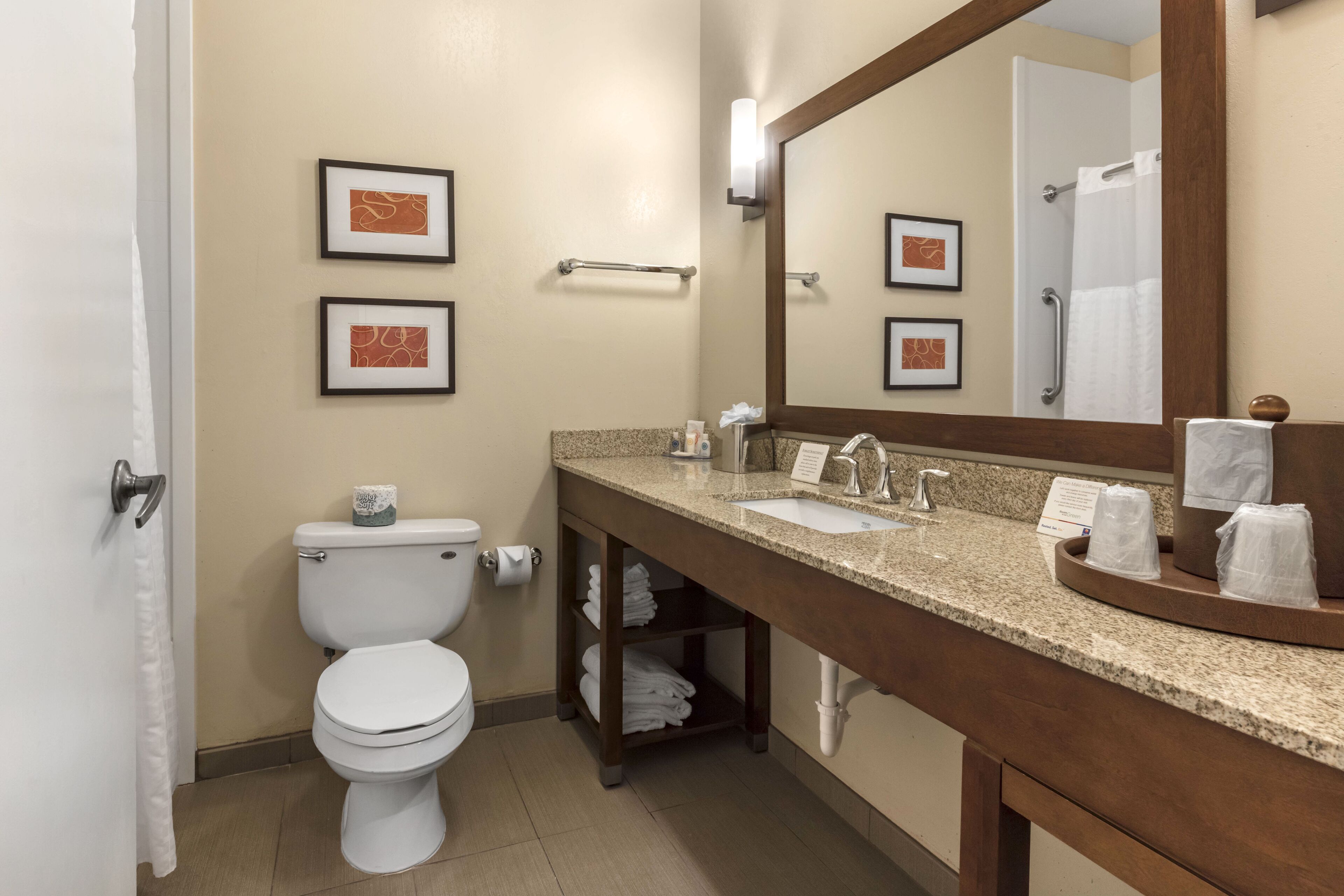 A modern bathroom with a toilet, sink, and mirror. There are framed pictures on the wall, a towel rack, and a shelf with folded towels.