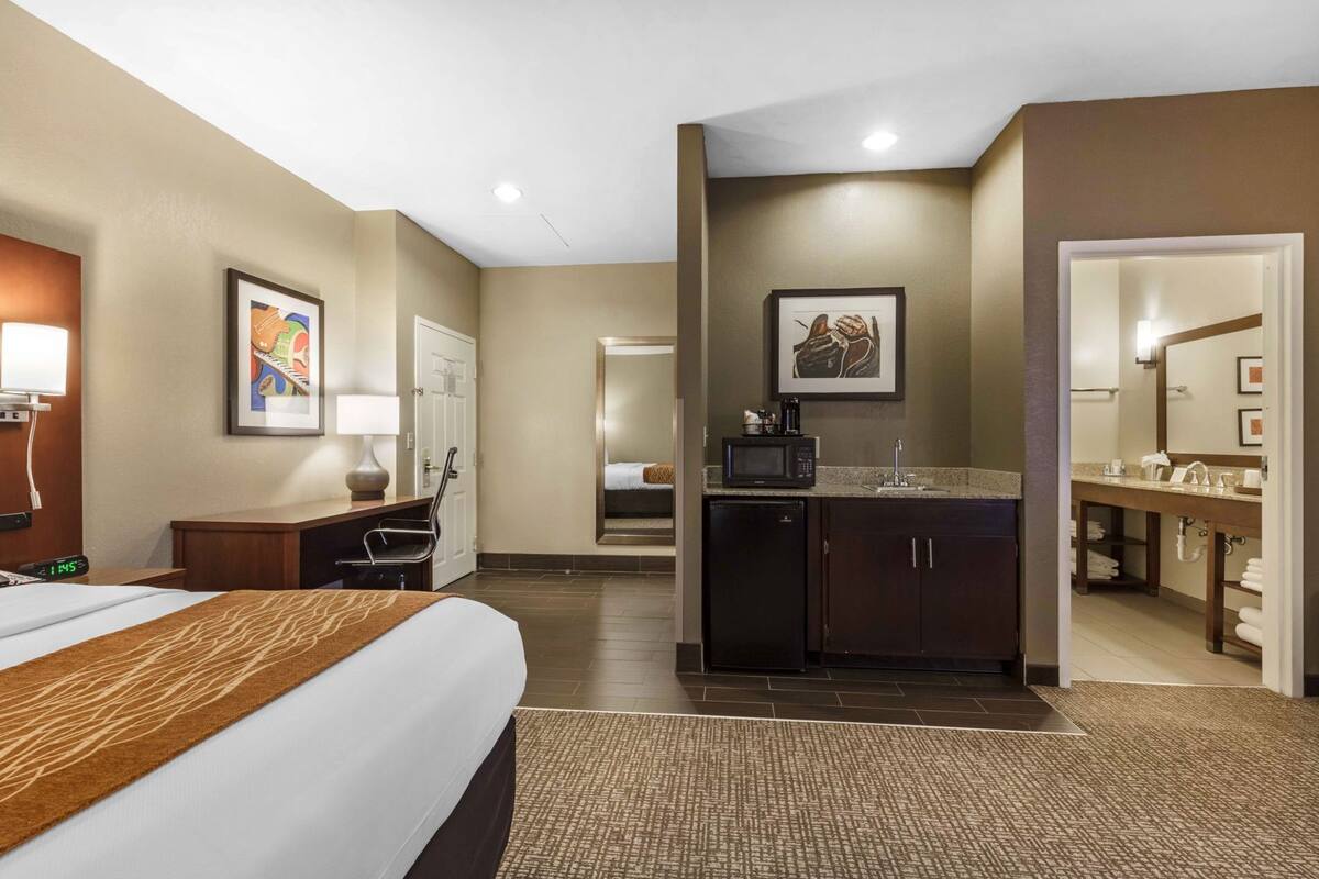A hotel room with a bed, desk, chair, kitchenette, and a bathroom.