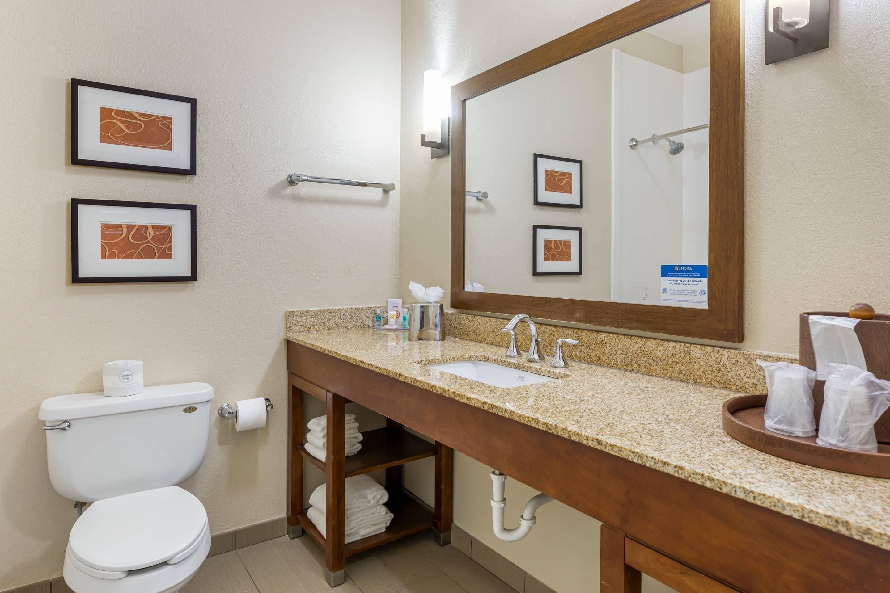 A hotel bathroom with a toilet, sink, and mirror. There are framed pictures on the wall, a towel rack, and a sign indicating housekeeping services.