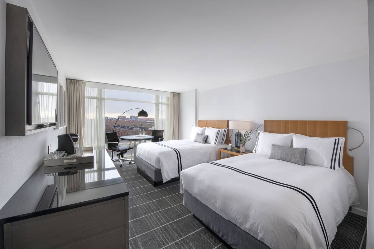 A hotel room with two beds, a desk, a chair, a large window with a city view, and a mirror.