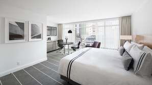 A hotel room with a large bed, a desk with a chair, a small table with a chair, and a view of buildings through large windows.