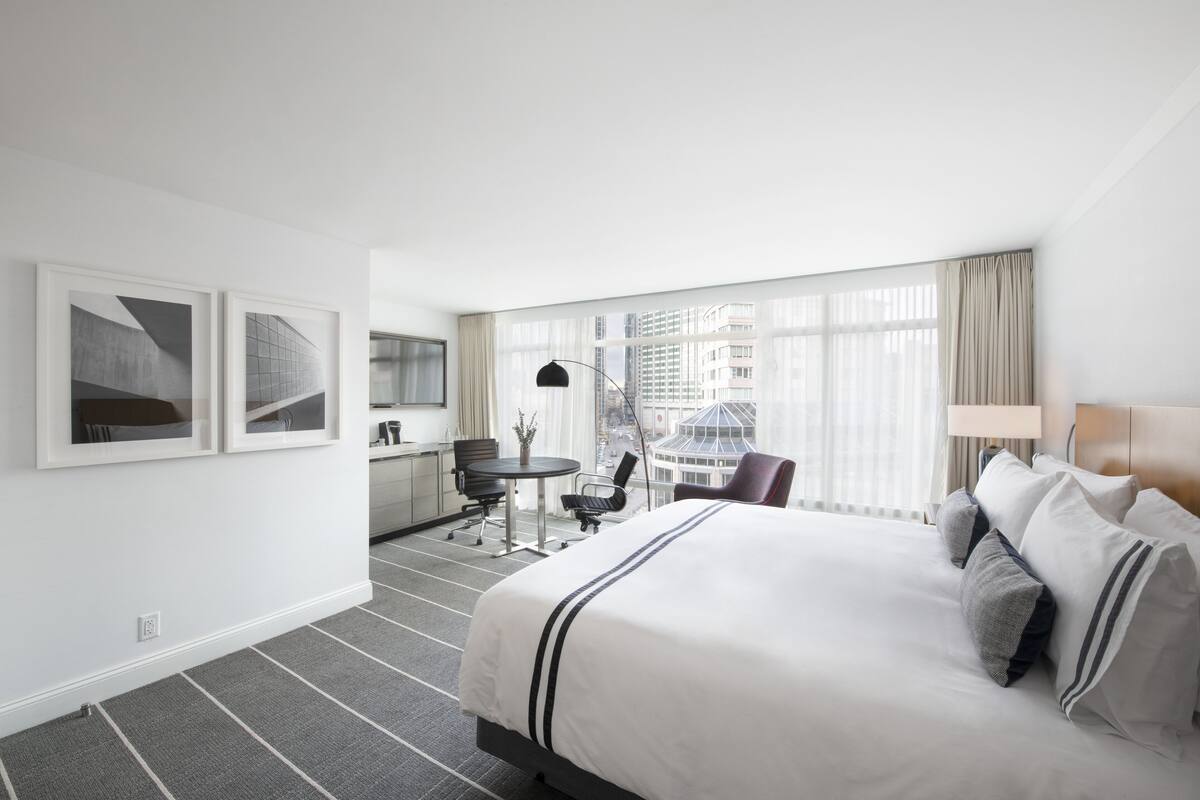 A hotel room with a large bed, a desk with a chair, a small table with a chair, and a view of buildings through large windows.