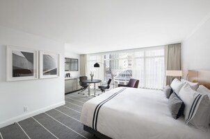 A hotel room with a large bed, a desk with a chair, a small table with a chair, and a view of buildings through large windows.