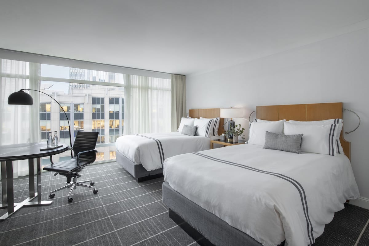 A hotel room with two beds, a desk, and a chair. There is a large window with a view of buildings.