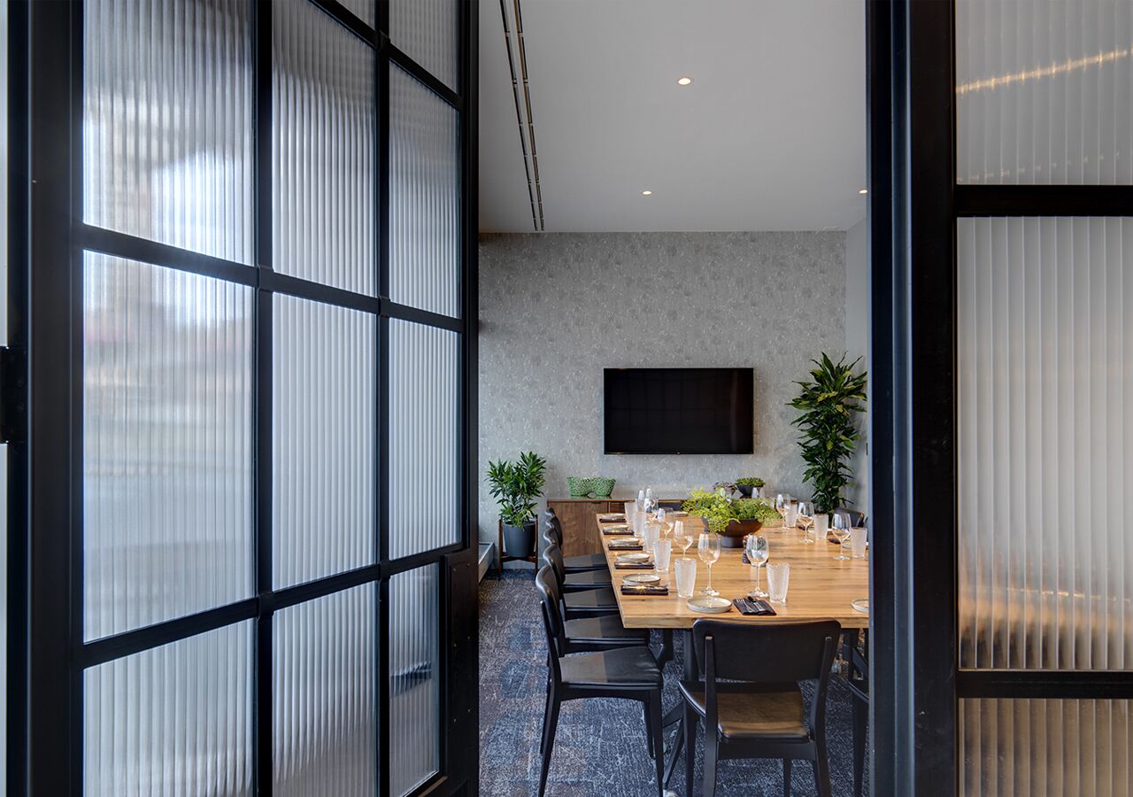 A modern conference room with a long table set for a meeting, large windows with blinds, and a flat-screen TV mounted on the wall.