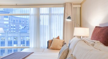 Een hotelkamer met een groot raam, een bed met veel kussens, een bedlampje en uitzicht op een stadsbeeld.