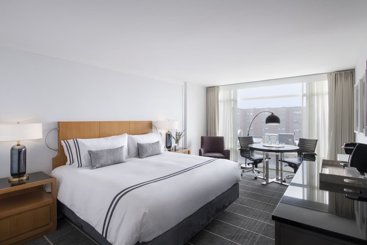 A hotel room with a large bed, a desk, and a chair. There is a window with curtains and a view of buildings outside.