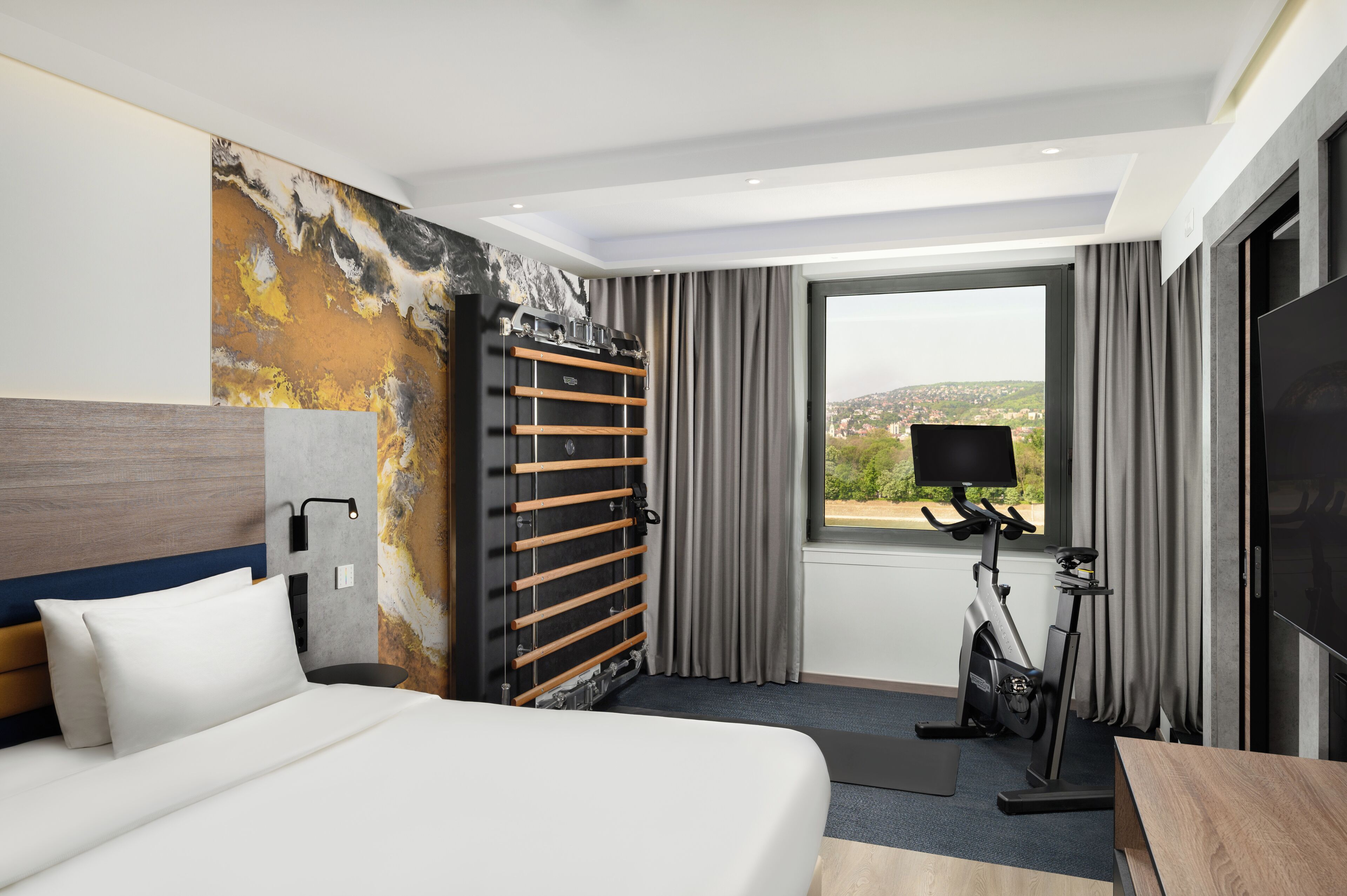 A hotel room with a bed, exercise bike, and a large window offering a city view.