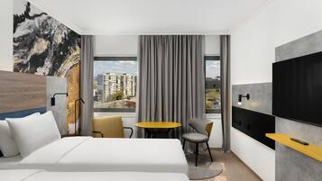 A modern hotel room with a large bed, a small round table, two chairs, a TV mounted on the wall, and a large window with curtains.