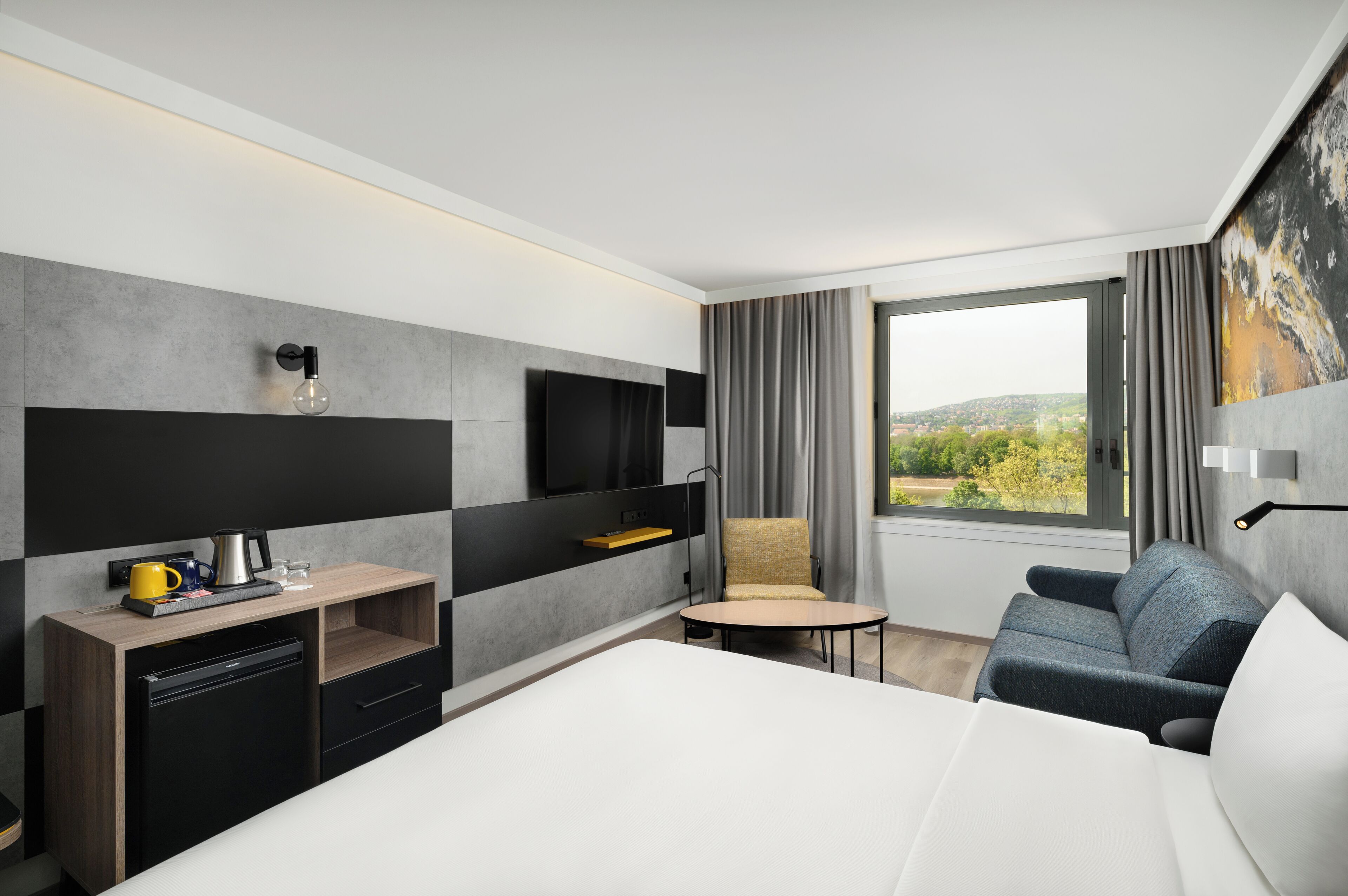 Habitación de hotel moderna con cama, escritorio con cafetera, silla, televisión y vista al exterior.