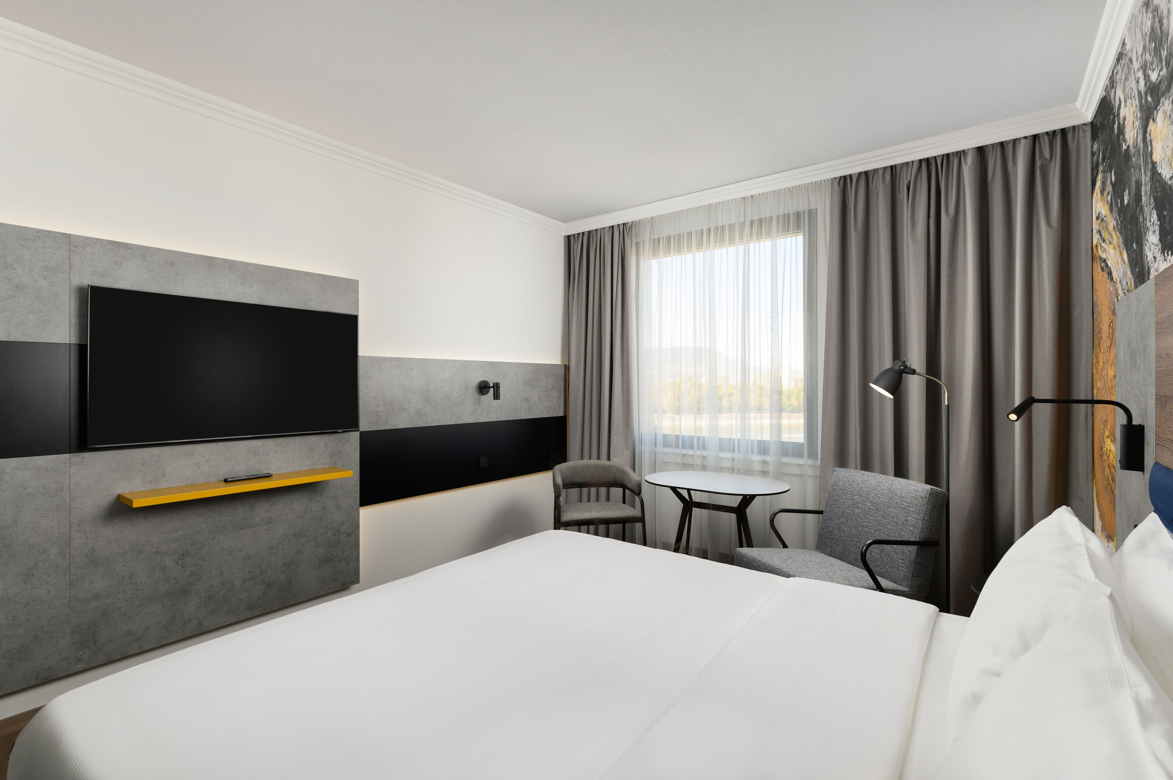Moderni hotellihuone, jossa on suuri sänky, harmaaseen seinään asennettu litteänäyttöinen televisio, pieni pöytä kahdella tuolilla sekä suuri ikkuna harmailla verhoilla.