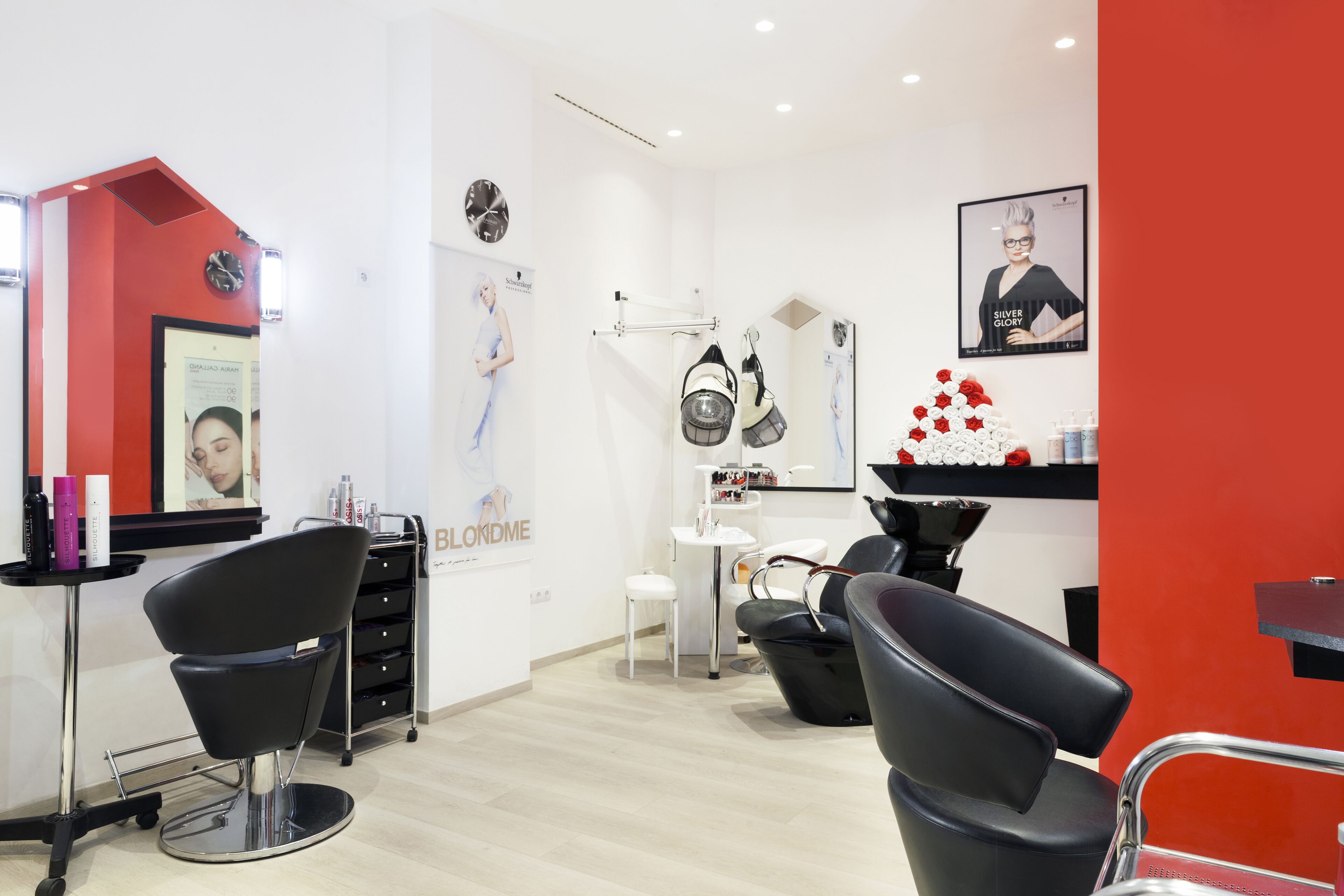 Um salão moderno com cadeiras pretas, uma parede de destaque vermelha e diversos produtos para cabelo em prateleiras.