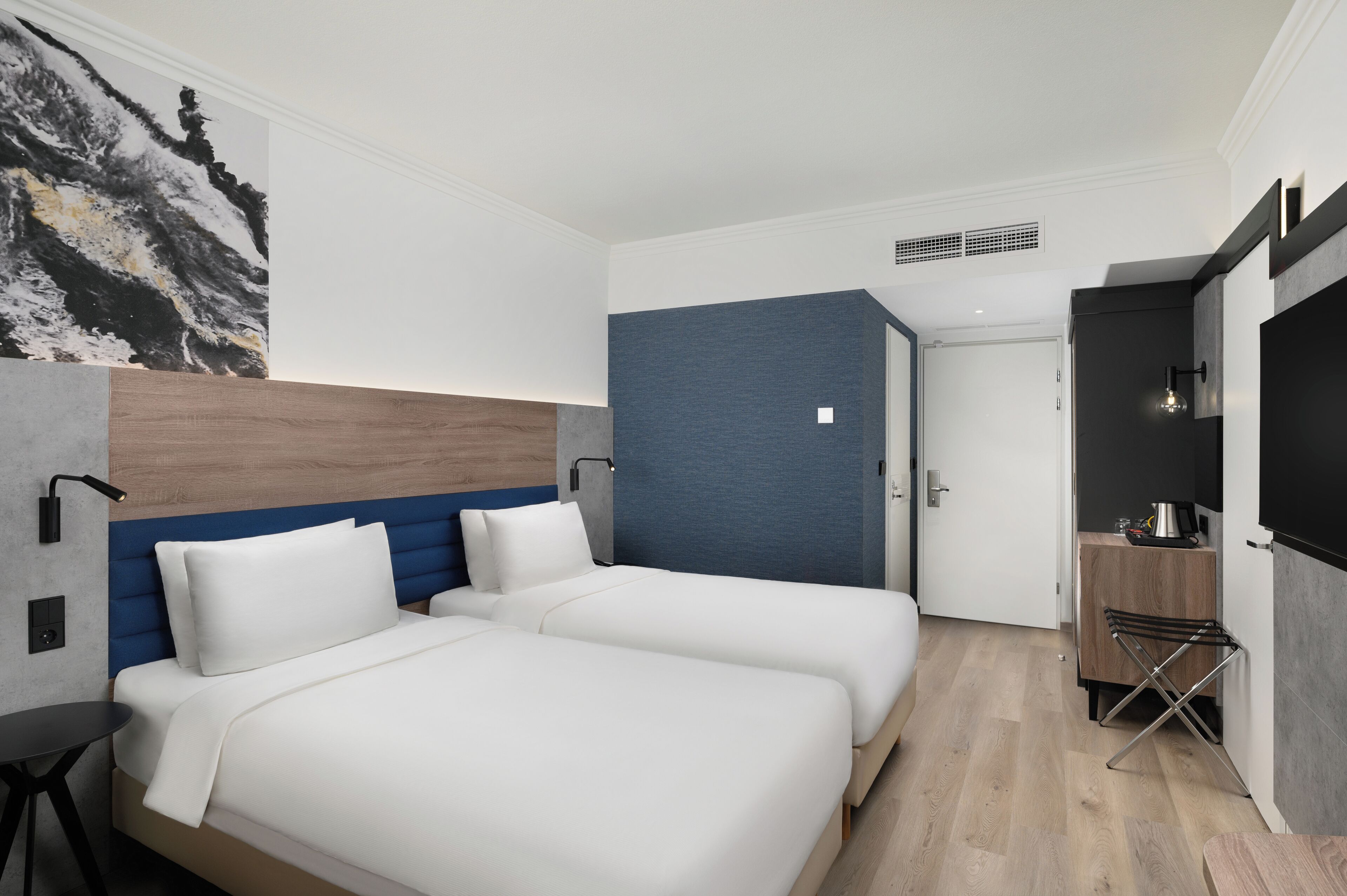 A modern hotel room with two beds, a wooden headboard, a blue accent wall, a small kitchenette, and a large artwork on the wall.