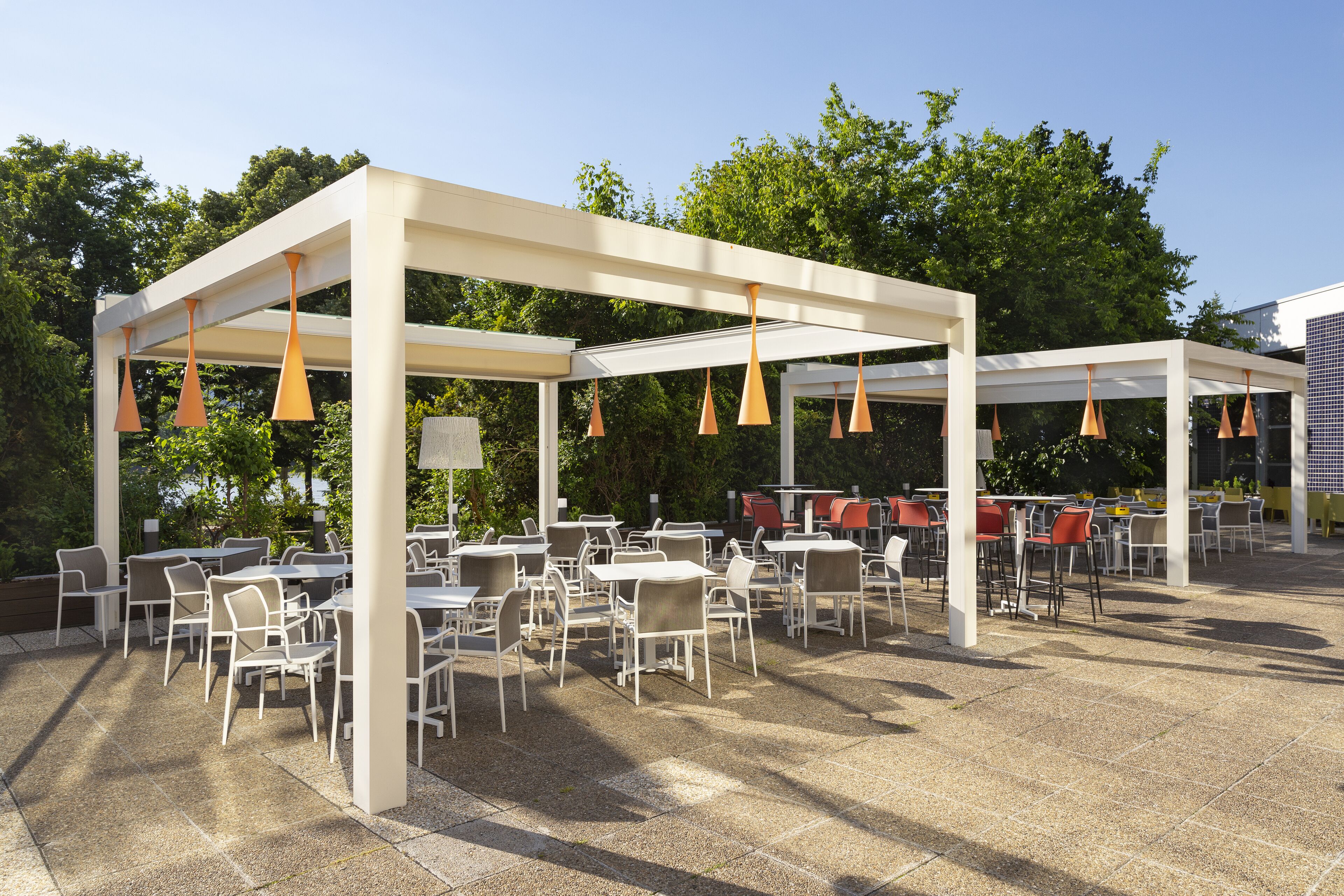 Un patio al aire libre con mesas y sillas, cubierto por una pérgola blanca con lámparas colgantes naranjas.
