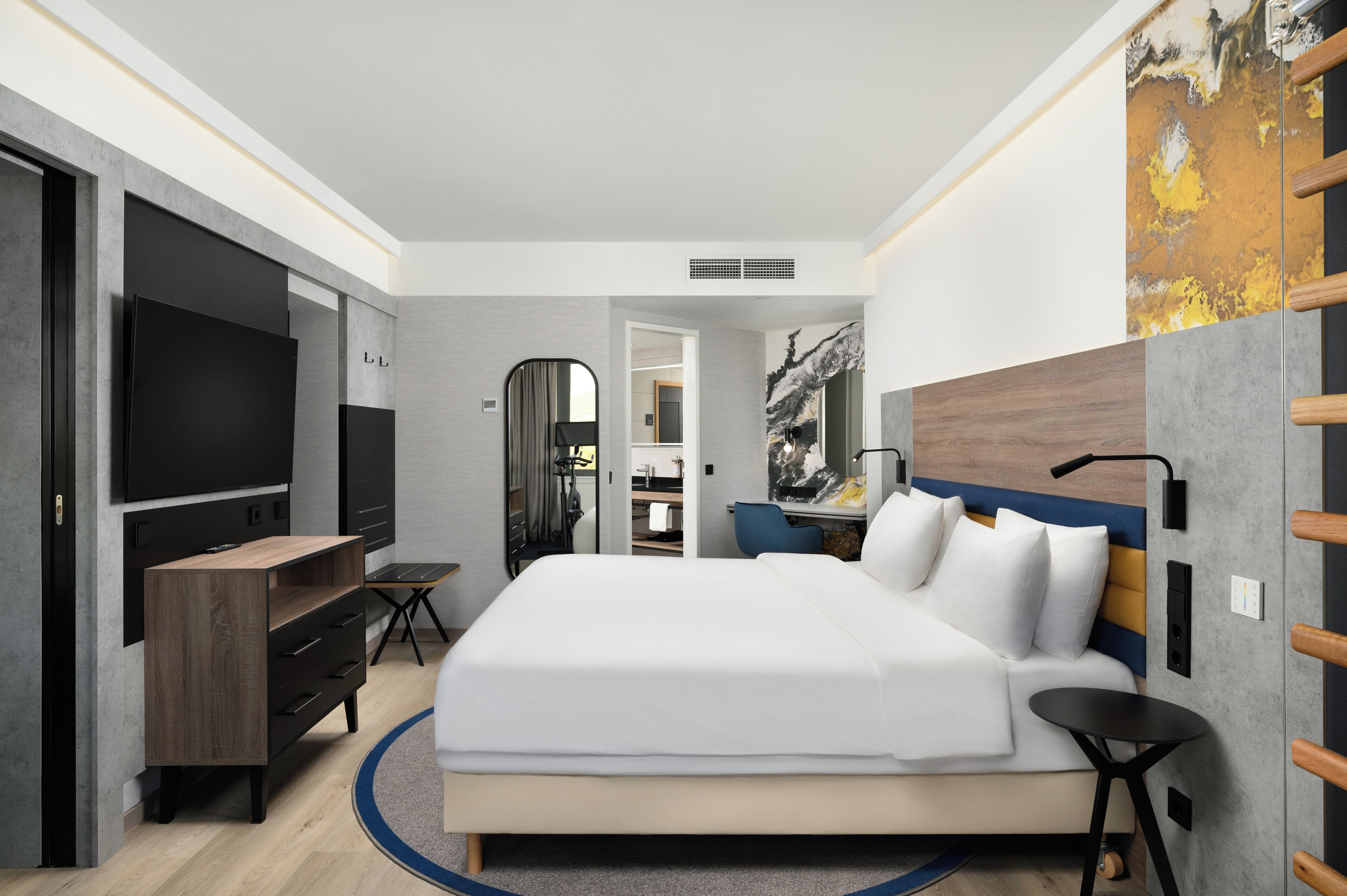 A modern hotel room with a large bed, a flat-screen TV, a desk, and a mirror.
