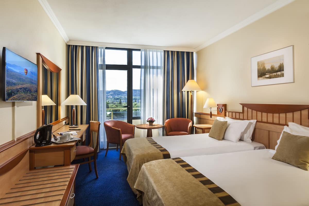 A hotel room with two beds, a desk, a chair, a TV, and a view of the outdoors.