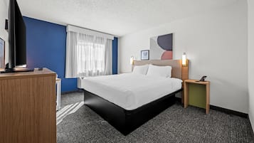 Et hotelværelse med en seng, et skrivebord, et fjernsyn og et natbord med en lampe.