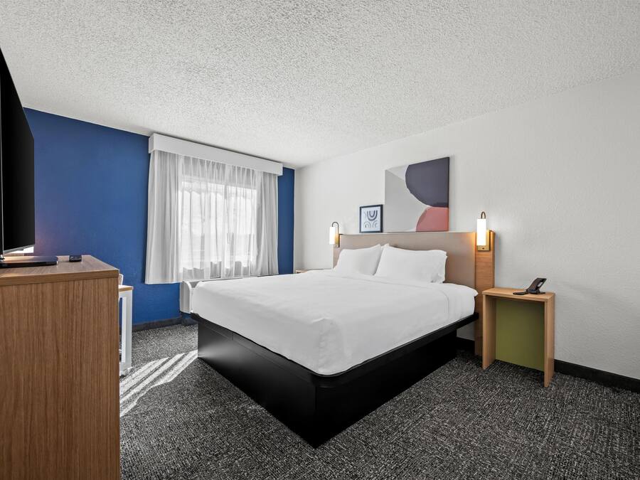 Et hotelværelse med en seng, et skrivebord, et fjernsyn og et natbord med en lampe.