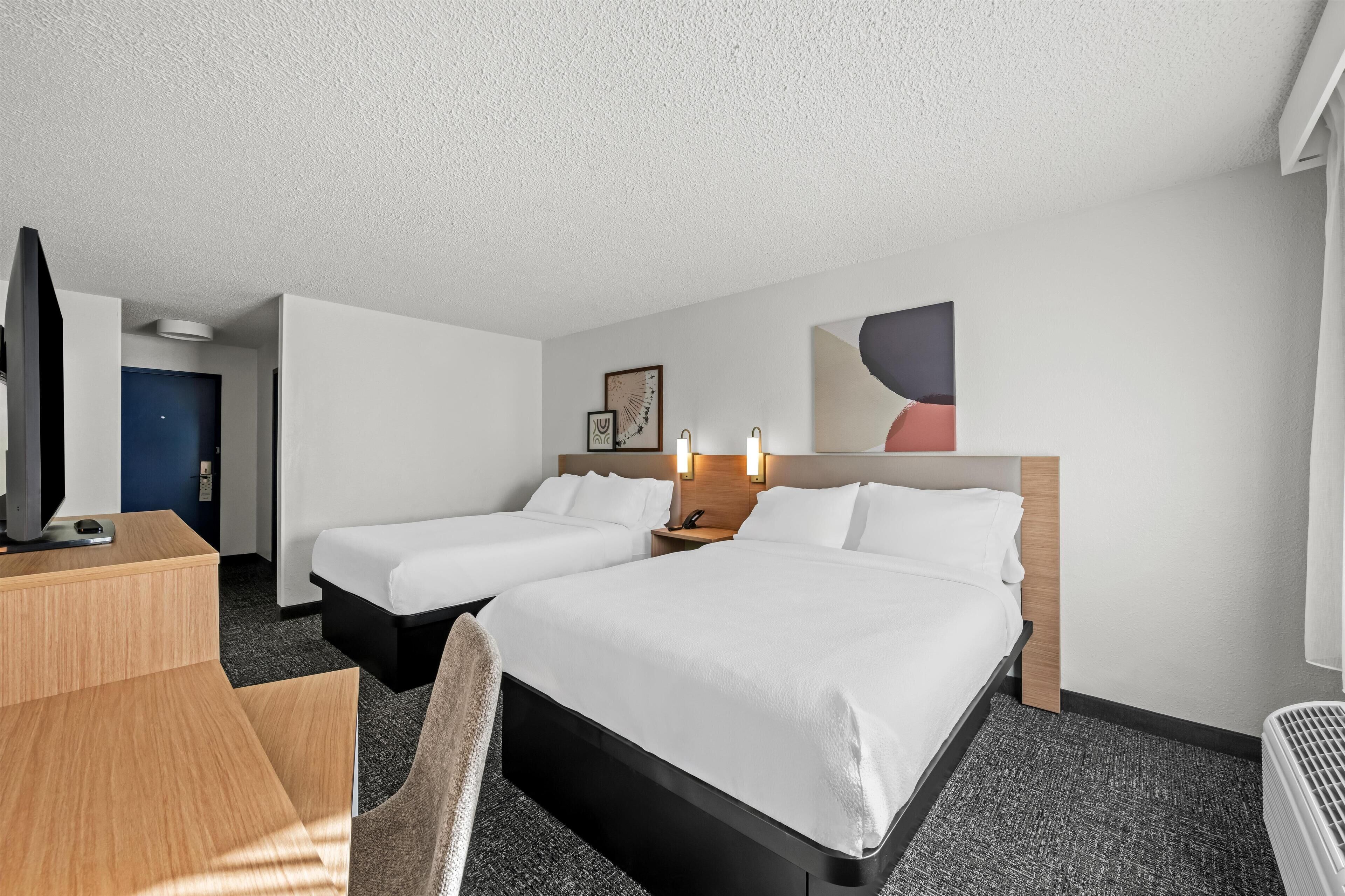 A hotel room with two beds, a desk, a chair, a TV, and a wall-mounted heater.