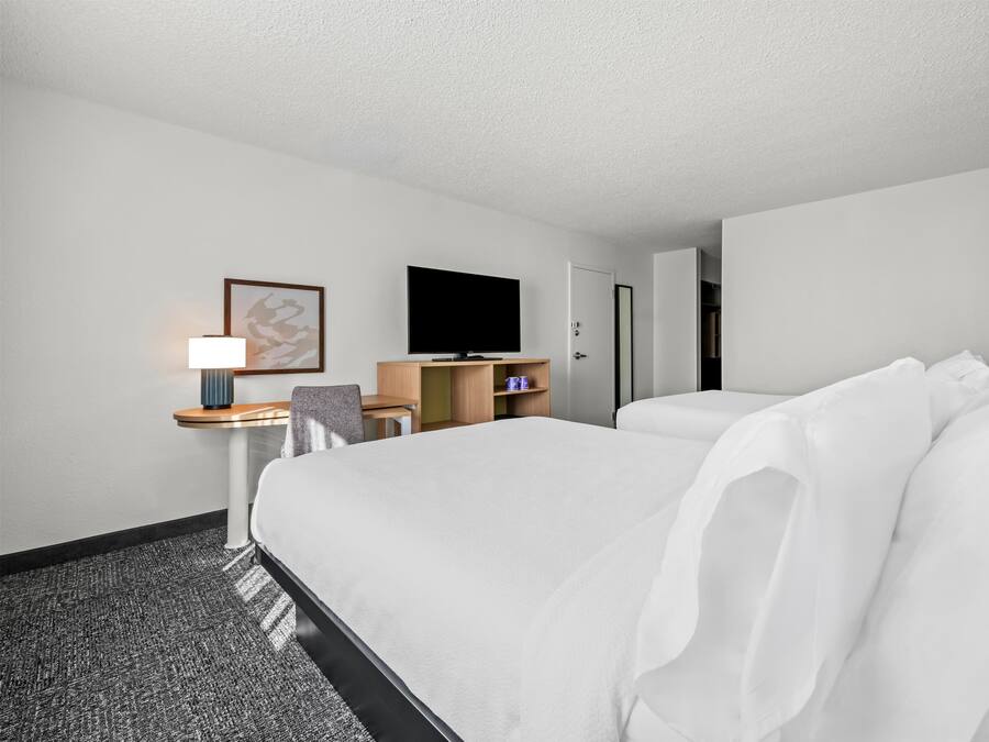 Quarto de hotel com uma cama grande, uma escrivaninha com luminária, televisão e guarda-roupa.