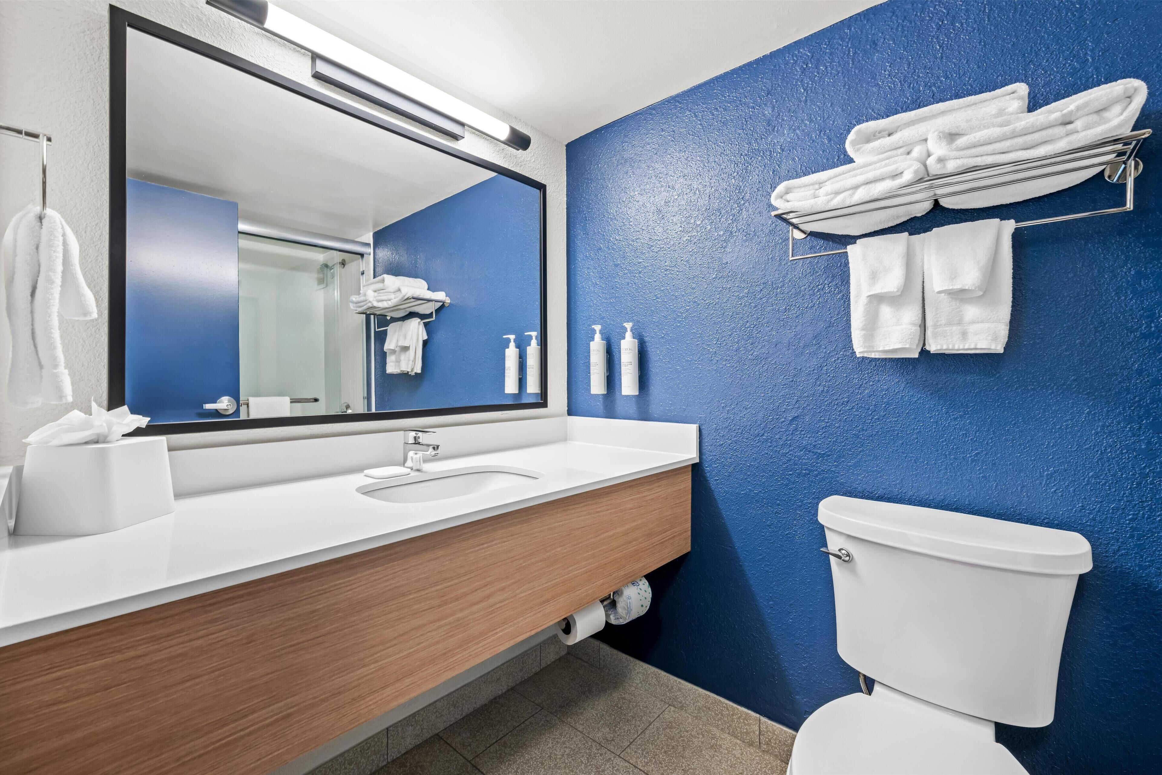 Un baño moderno con una pared azul de acento, un mueble de tocador de madera, un lavabo blanco, un inodoro y un perchero con toallas dobladas.