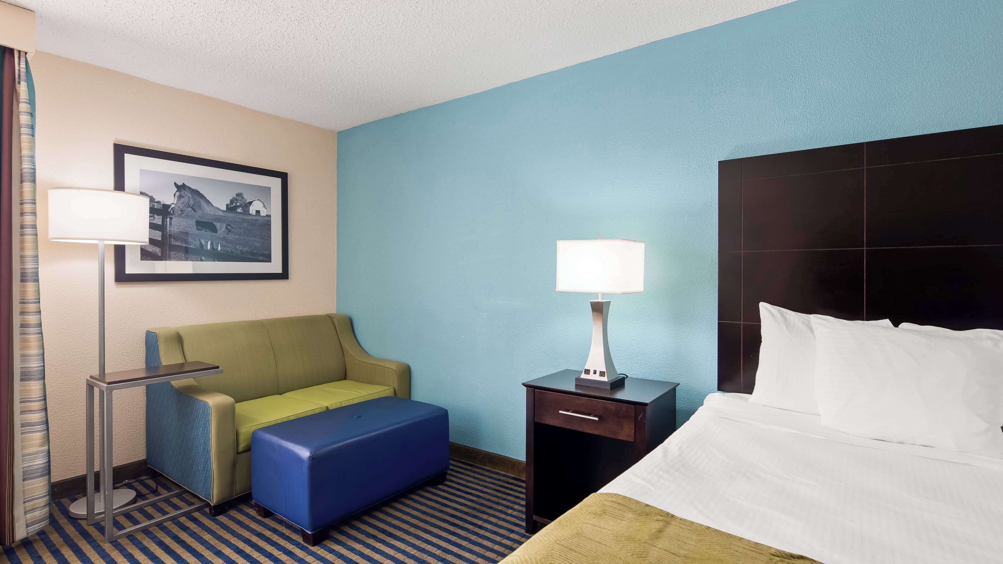 Habitación de hotel con cama, escritorio con lámpara, sofá, mesita pequeña y un cuadro en la pared.