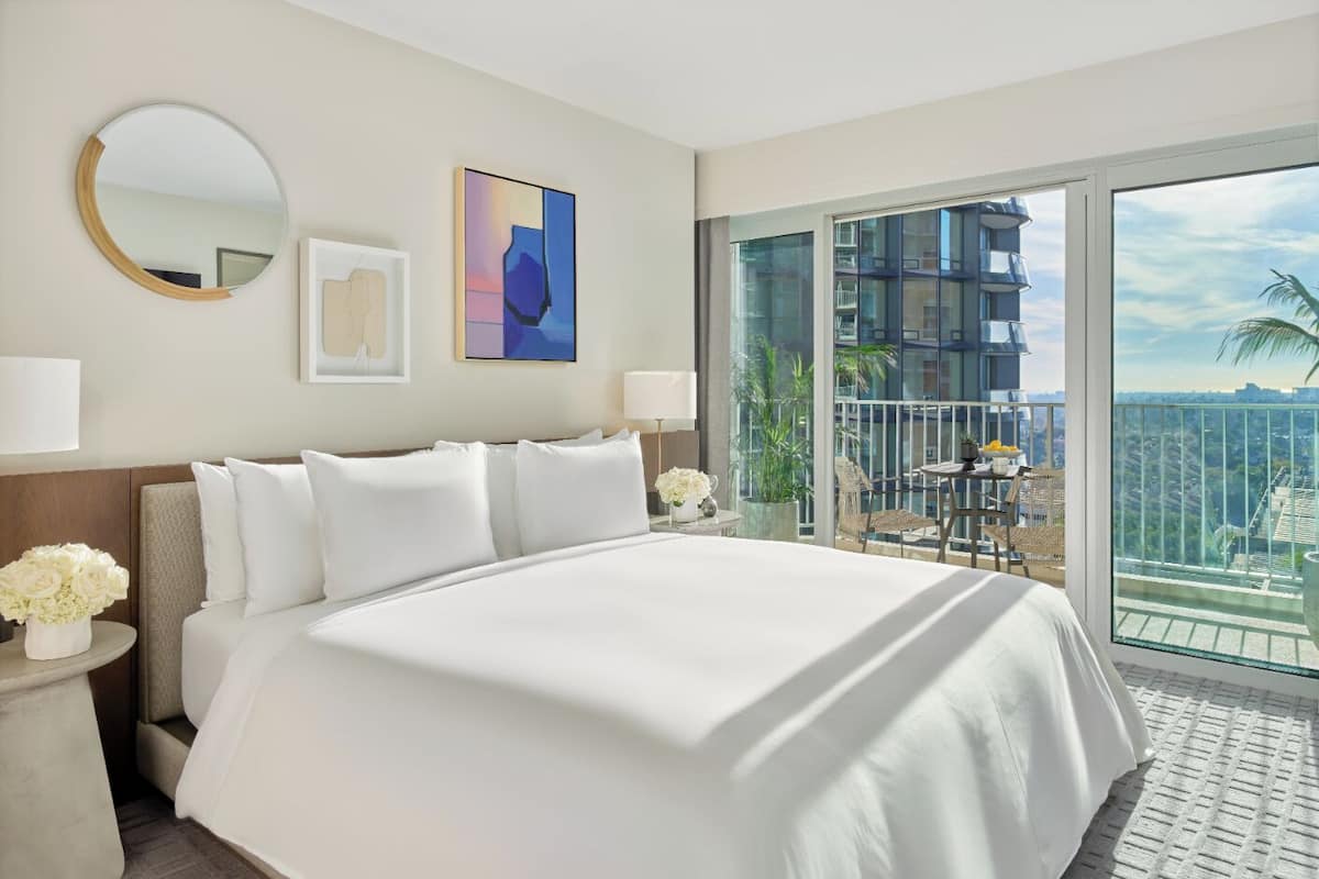 Ein Hotelzimmer mit einem großen Bett, einem runden Spiegel, einem gerahmten abstrakten Gemälde und einem Balkon mit Blick auf eine Stadtlandschaft.