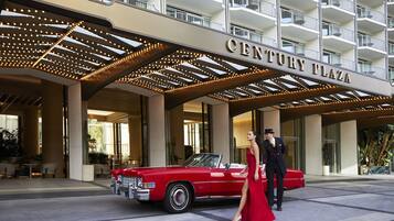 En röd, klassisk bil parkerad framför hotellet Century Plaza, med en man och en kvinna som bär formella kläder.