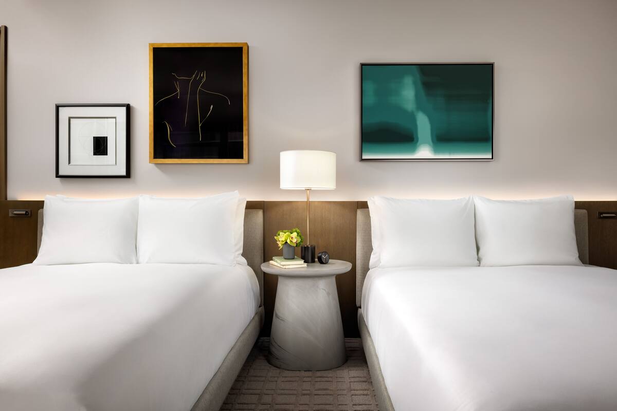 Ein Hotelzimmer mit zwei Betten, einem runden Beistelltisch mit Lampe, gerahmten Bildern und einer kleinen Vase mit Blumen.